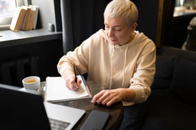 elderly-woman-drinking-coffee-at-cafe-while-working-on-her-laptop-and-writing-on-her-notebook