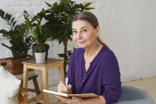 portrait-of-busy-modern-50-yeard-old-middle-aged-woman-in-violet-shirt-writing-in-copybook-making-plans-looking-with-positive-friendly-smile-sitting-on-chair-surrounded-with-green-plants